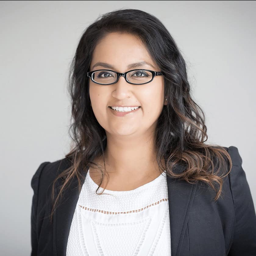 A woman with long dark hair, wearing glasses, a white blouse, and a black blazer, smiles at the camera against a plain light gray background, representing the Academy for Sustainable Innovation.