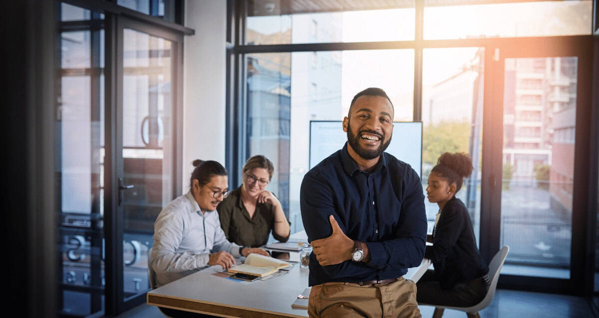 A man in a dark shirt stands smiling with arms crossed, leaning against a desk. Behind him, four colleagues, modern-day practitioners of their craft, sit at a table engaged in discussion and working on documents. The glass-walled office boasts a sunny outdoor view, symbolizing the bright possibilities of 2023.