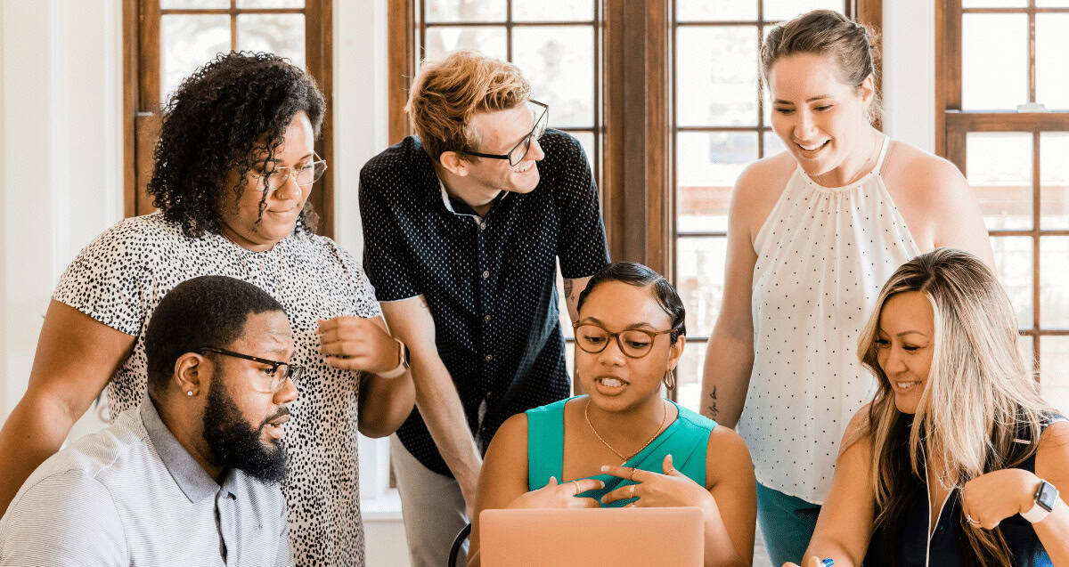 A group of six diverse individuals, three men and three women, are gathered around a laptop, engaged in discussion. They are smiling and appear collaborative. The bright room with large windows highlights how the modern workforce is driven by values of teamwork and inclusivity.