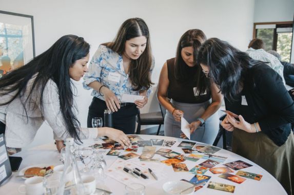 Four women stand around a table covered with various cards and papers, attentively examining and discussing the items. The setting appears to be a meeting or workshop in a bright, modern room.