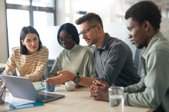 Four people sit at a table in an office, looking at a laptop screen. One man gestures while speaking, and the others listen attentively. Cups and glasses are on the table.