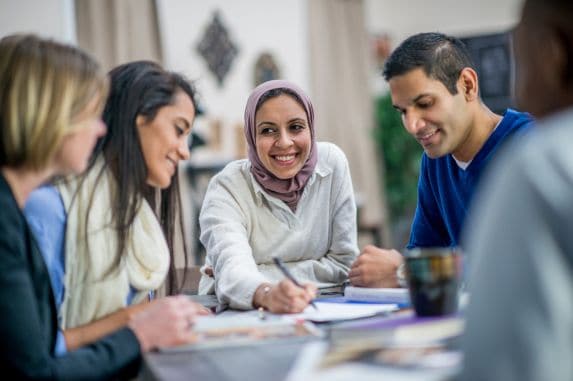 A group of four people sit around a table engaged in discussion. One woman wearing a hijab is smiling and writing, while the others listen attentively. Papers and notebooks are spread out on the table.