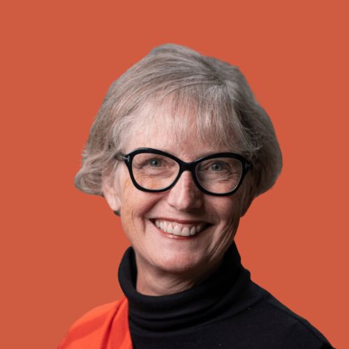 A smiling woman with short gray hair and black-framed glasses is pictured against an orange background. She is wearing a black top with an orange garment draped over one shoulder.