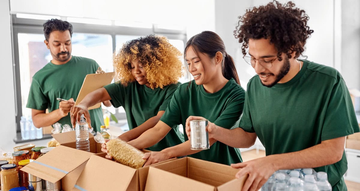 Four people in green shirts are packing food items like canned goods and pasta into cardboard boxes. Working together in a well-lit room with large windows, they are likely engaged in a community service activity organized by a climate action nonprofit.