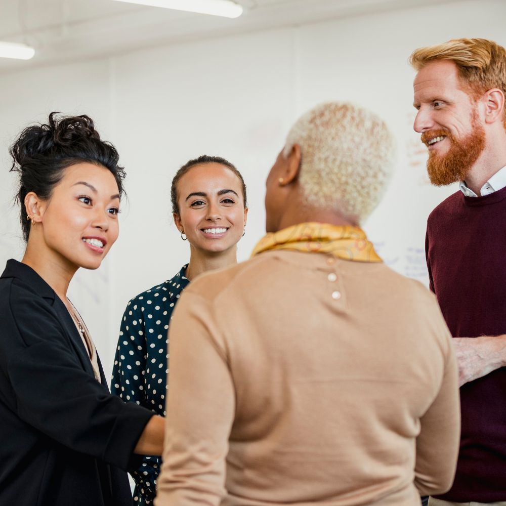 A diverse group of four people, three women and one man, are standing and having a friendly conversation in a well-lit office setting. Embracing collaboration, the woman with short blond hair and a beige top has her back to the camera, highlighting an environment of shared leadership.