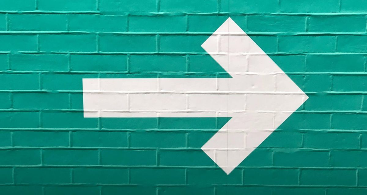 A large white right-pointing arrow is painted on a teal brick wall. The arrow spans multiple bricks, creating a clean, bold graphic against the colored background.
