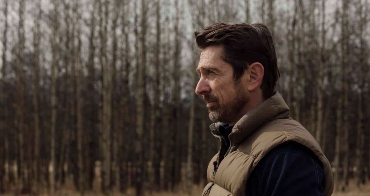 A man with short brown hair and a beard stands outdoors, gazing to the side. He is wearing a brown puffer vest over a dark shirt. Bare trees with no leaves fill the background, suggesting a cold or autumn day.