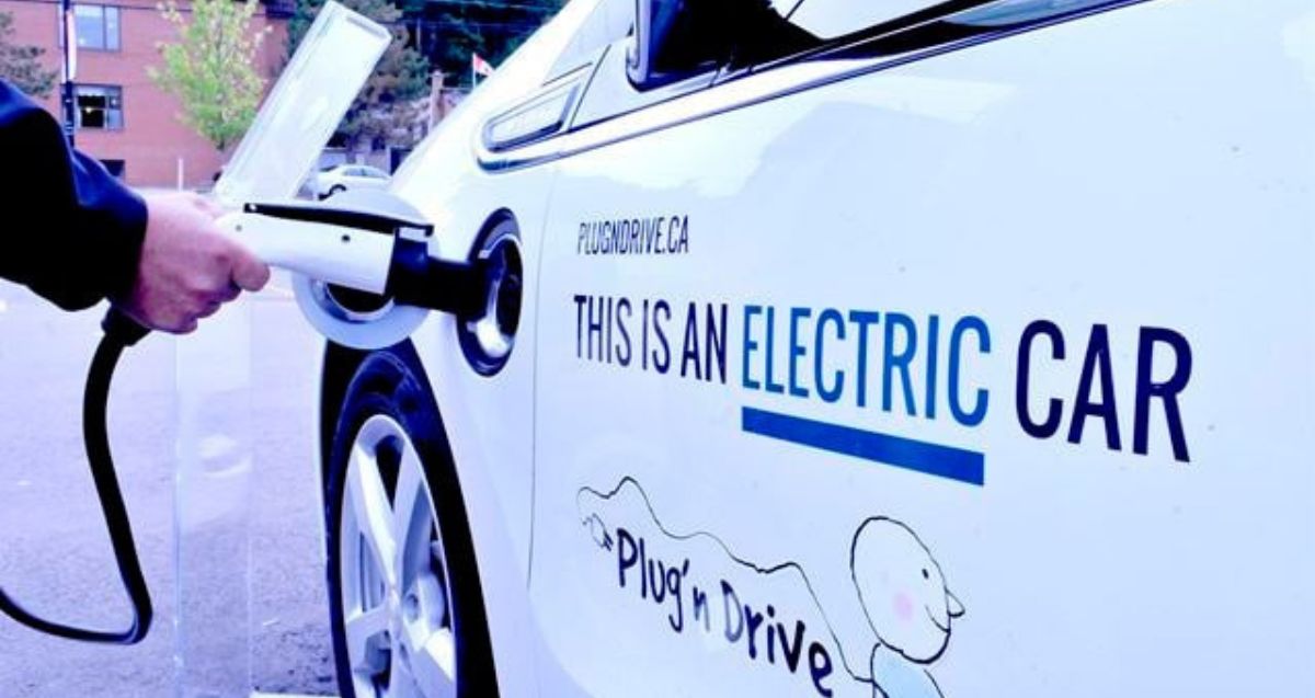 Person charging an electric car. The side of the white car displays text: "This is an electric car" and "Plug'n Drive" under an illustration of a plug with a cheerful face. The background features a building and trees.