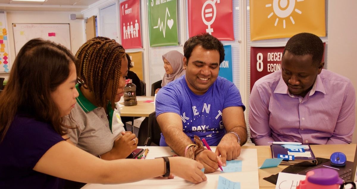 A diverse group of four people sit around a table, engaging in a collaborative activity. They are smiling and appear to be writing on colorful sticky notes. The background features banners with various symbols and numbers, likely representing different goals or themes.