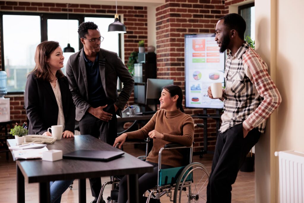 Four colleagues are gathered in an office, engaged in conversation. One person is seated in a wheelchair, and three others are standing. A large screen displaying graphs is in the background. The office has exposed brick walls and large windows.