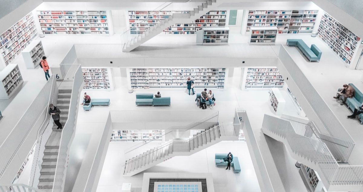 A spacious, modern library with multiple levels connected by staircases. The white interior is accented by teal seating areas. People are walking, reading, or sitting. Shelves filled with books line the walls on each level. Bright lighting enhances the airy atmosphere.