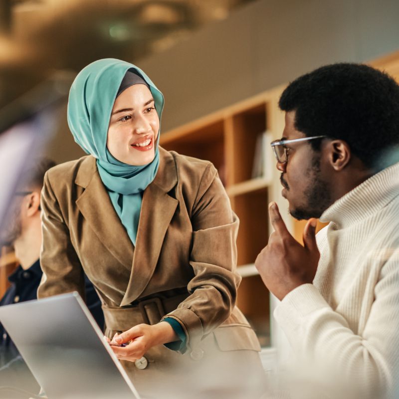 A woman wearing a teal hijab and beige blazer is smiling and discussing climate initiatives with a man wearing glasses and a white turtleneck sweater. The man is seated, gesturing with his hand. They are in an office setting with bookshelves in the background.