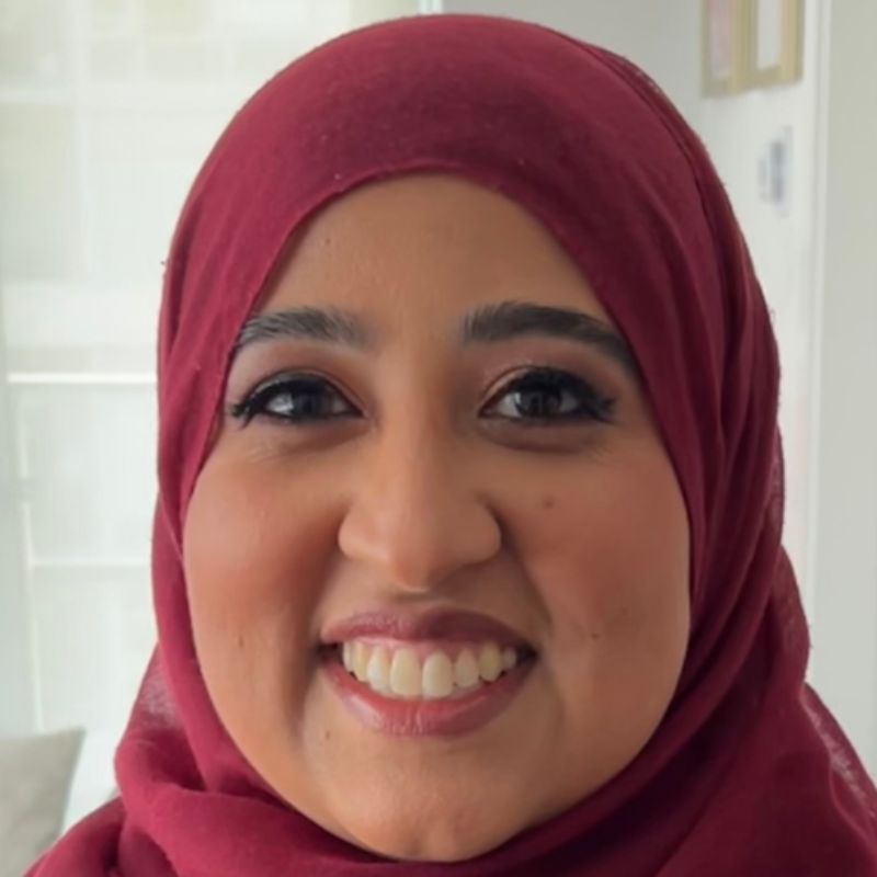 A woman wearing a maroon hijab smiles warmly at the camera. She has dark eyes, and her makeup includes pink eyeshadow and lipstick. The background is softly lit and out of focus, suggesting an indoor setting.