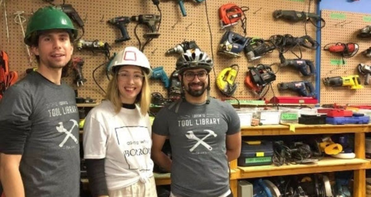 Three people in safety helmets posing together in a workshop filled with various tools hanging on the wall. The person on the left has a green helmet, the person in the middle has a white helmet, and the person on the right has a bike helmet and glasses.