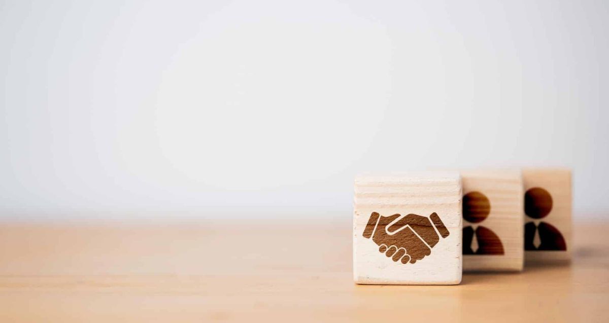 Three wooden blocks on a table, with the block in the foreground showing an image of a handshake. The two blocks in the background display icons of people. The background is light and out of focus, emphasizing the blocks.