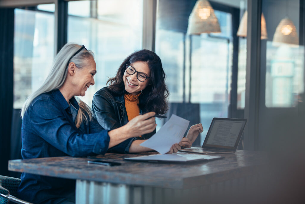 Two women, one with gray hair and one with dark hair, smiling and reviewing documents at a table with laptops in a modern office setting.