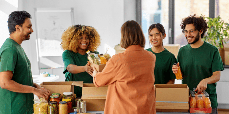 A group of people wearing green shirts are smiling and packing food items into cardboard boxes at a nonprofit community center. A woman in an orange shirt, one of the leaders, stands with them, holding a box. The scene is vibrant and filled with a spirit of giving.