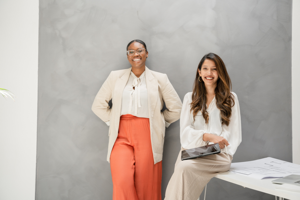 Two women are smiling in an office setting. One wears glasses, a beige blazer, and orange pants and stands with hands behind her back. The other, with long hair, wears a white blouse and tan pants and is seated with a tablet on her lap. They stand in front of a gray wall.