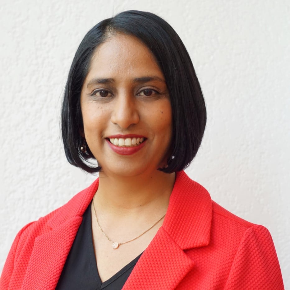 Priya Bala Miller, with short black hair, smiles at the camera. She is wearing a red blazer over a black top and a delicate necklace. The background is a plain white wall.