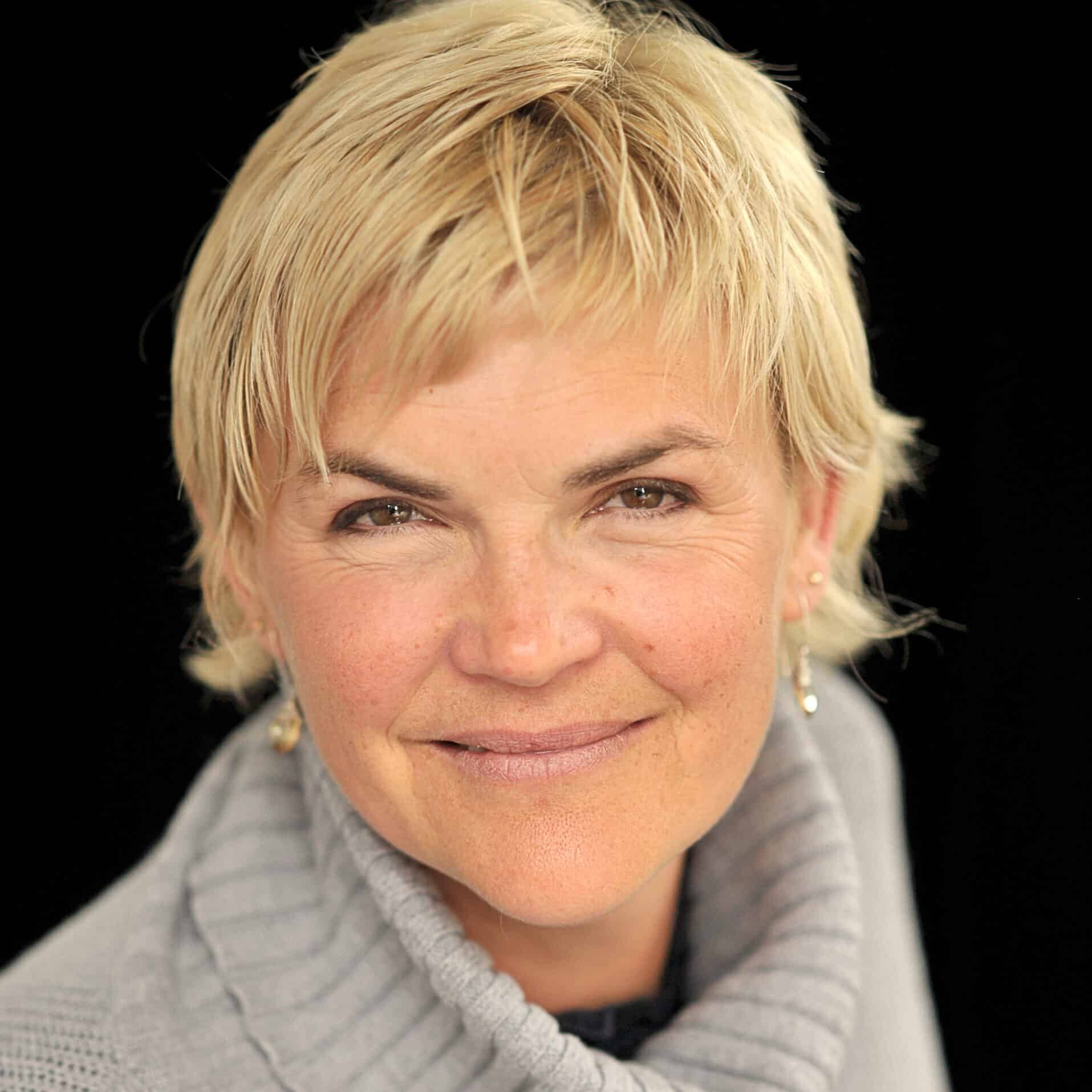 Anne-Marie Daniel, a woman with short, blonde hair, smiles gently at the camera. She has light skin and is wearing a gray, ribbed turtleneck sweater. The background is black, highlighting her face and friendly expression.