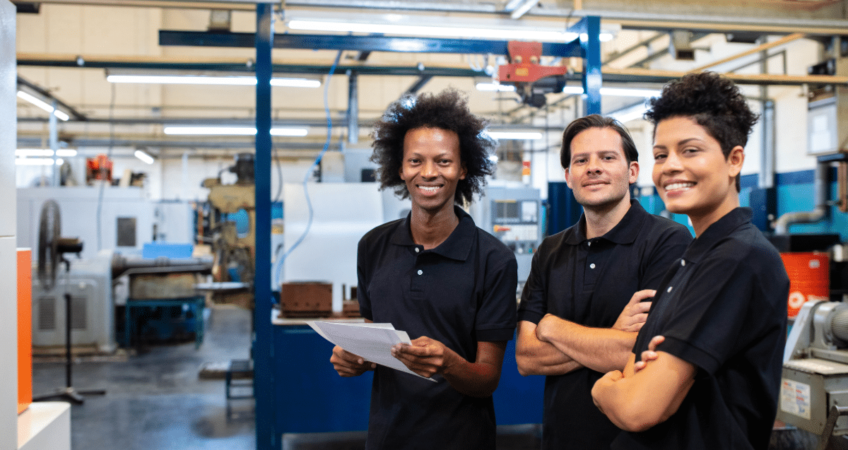 Three individuals wearing black polo shirts stand in a brightly-lit factory, smiling at the camera. One person holds papers, while the other two have their arms crossed. Machinery and equipment are visible in the background, highlighting their dedication to bridging the green skills gap in manufacturing.