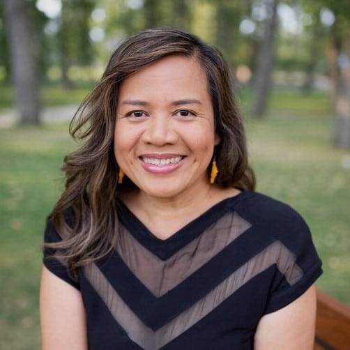 A person with long hair and a warm smile is sitting outdoors on a wooden bench. They are wearing a black dress with a sheer, V-shaped detail at the neckline. The background features green grass and blurred trees, indicating a park or natural setting—a serene moment reminiscent of Marie Jurcevic's elegance.