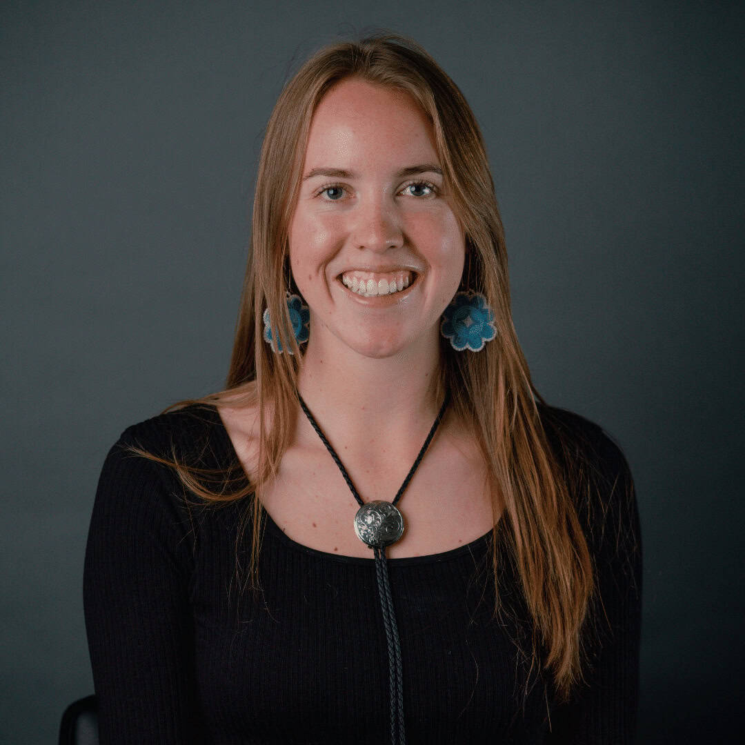 Smiling woman with long brown hair wearing a black top and blue flower-shaped earrings, reminiscent of a Freddie Huppé Campbell portrait, set against a dark background.