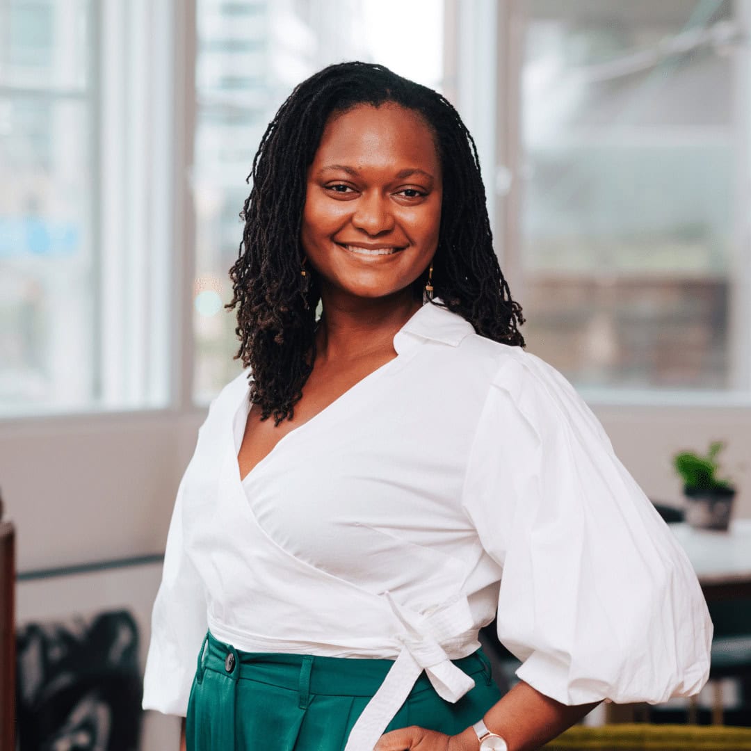 Alicia Richins, a woman with long curly hair wearing a white wrap blouse and green pants, stands smiling in a bright, modern office with large windows and plants in the background.