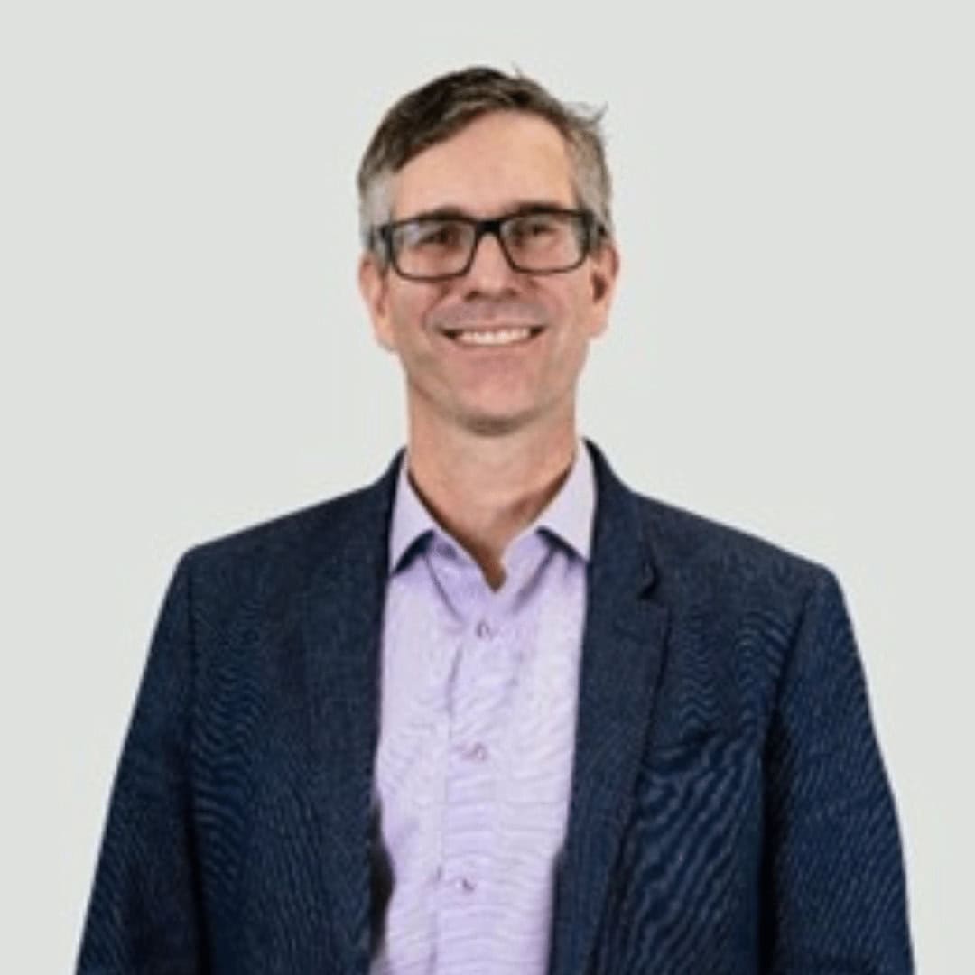 A man with short brown hair and glasses, wearing a dark blazer over a light purple button-up shirt, smiles at the camera against a plain light background—reflecting the spirit of Building Trust in every interaction.