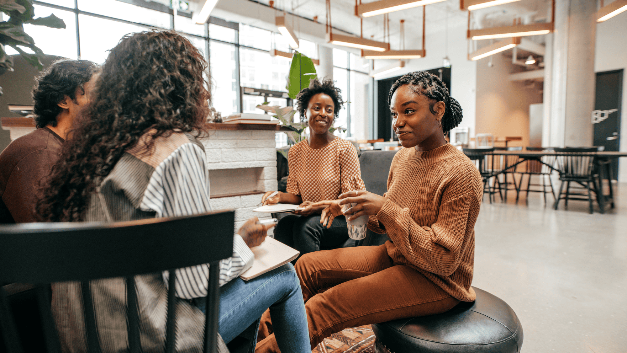 Four people sit together in a modern, bright workspace, engaged in conversation about leadership. Two women with curly hair face the camera, one holding a notebook and another a glass of water, all appearing relaxed and friendly.