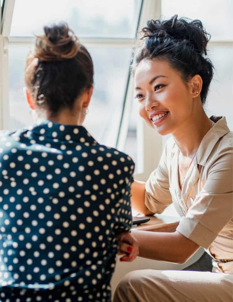 Two women sit across from each other at a table by a window. One woman, smiling, faces the camera, while the other has her back to it. Their friendly conversation suggests a discussion on leadership or climate action.