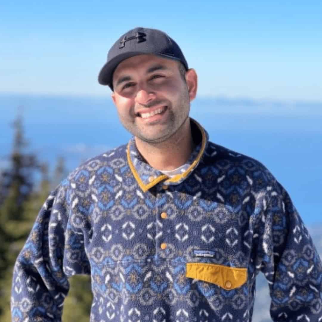 Yousef Sadat, a smiling man in a patterned blue fleece and dark baseball cap, stands outdoors with trees and a clear blue sky in the background.