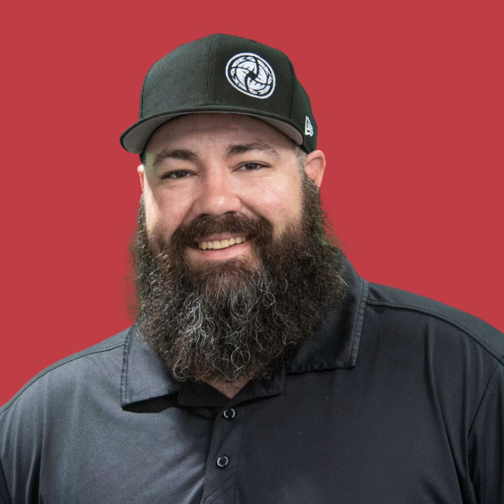 A bearded man wearing a black baseball cap with a logo and a black collared shirt, smiling against a solid red background, ready to discuss Let's Talk Leadership.