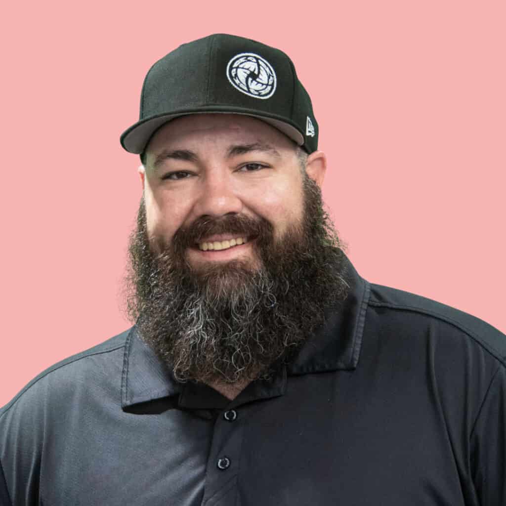 A bearded man wearing a black cap with a circular logo and a black collared shirt smiles in front of a pink background.