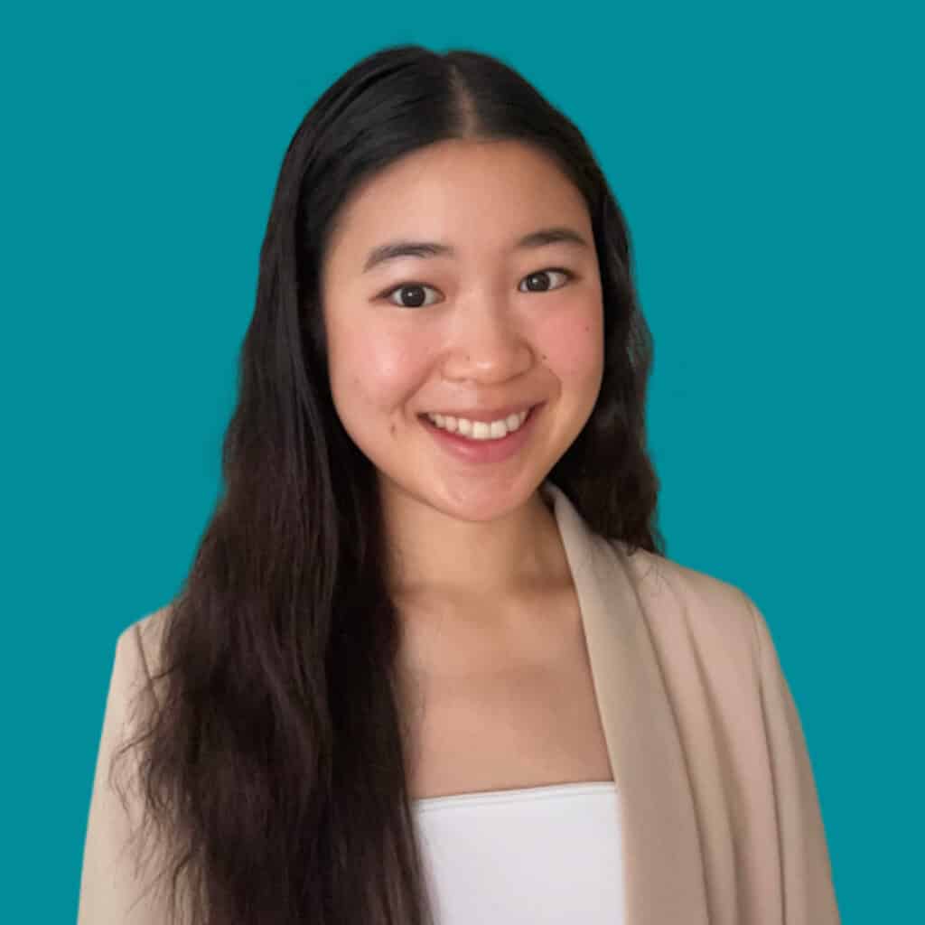 A young woman with long dark hair smiles at the camera. She is wearing a beige blazer over a white top, standing in front of a solid teal background.