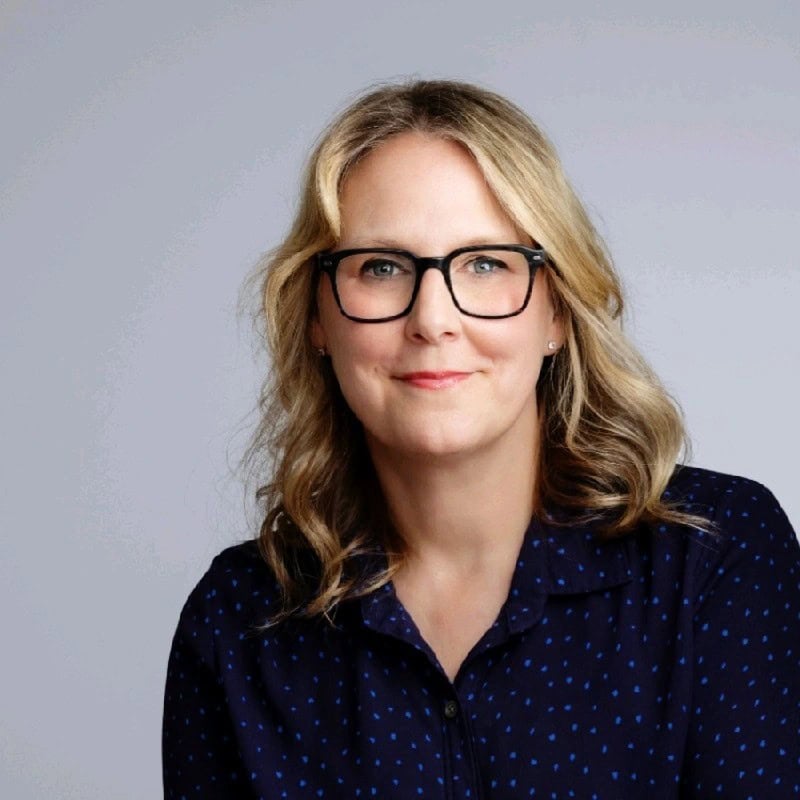 A woman with wavy blonde hair and glasses, wearing a navy blue shirt with small dots, smiles softly against a plain, light gray background—perfect for testimonials.