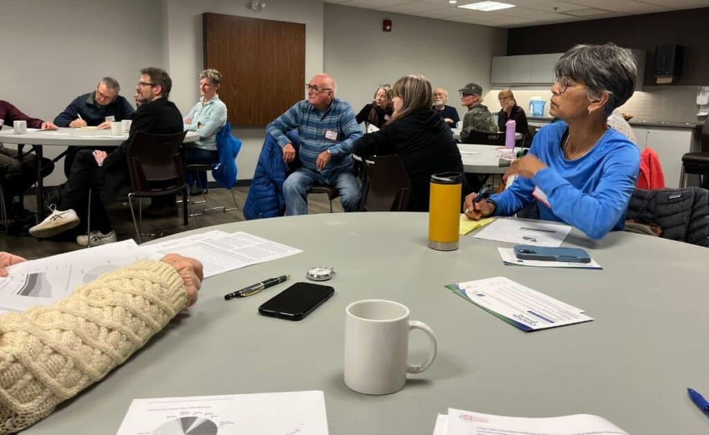 A group of adults sits around tables in a meeting room, listening attentively. Papers, pens, mugs, and phones are on the tables as they discuss custom engagements. The atmosphere appears focused and engaged.