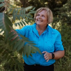 A smiling older woman wearing a bright blue shirt stands outdoors, gently touching the leaves of a tree or bush. Lush green foliage surrounds her in a sunlit natural setting.