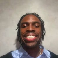 A person with dark skin and short dreadlocks smiles widely at the camera, wearing a blue and white striped collared shirt with a dark sweater.