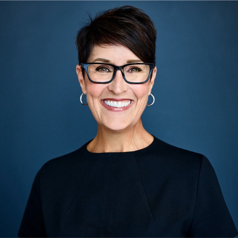 A woman with short dark hair, wearing glasses, silver hoop earrings, and a dark top, smiles warmly against a plain dark blue background—perfect for featuring client testimonials.