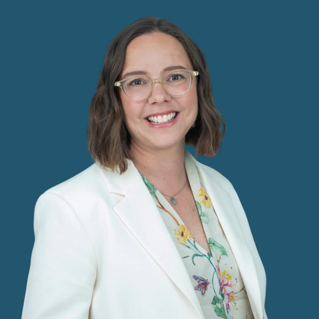 A woman with short brown hair and glasses smiles, wearing a white blazer over a floral top, standing in front of a solid teal background.