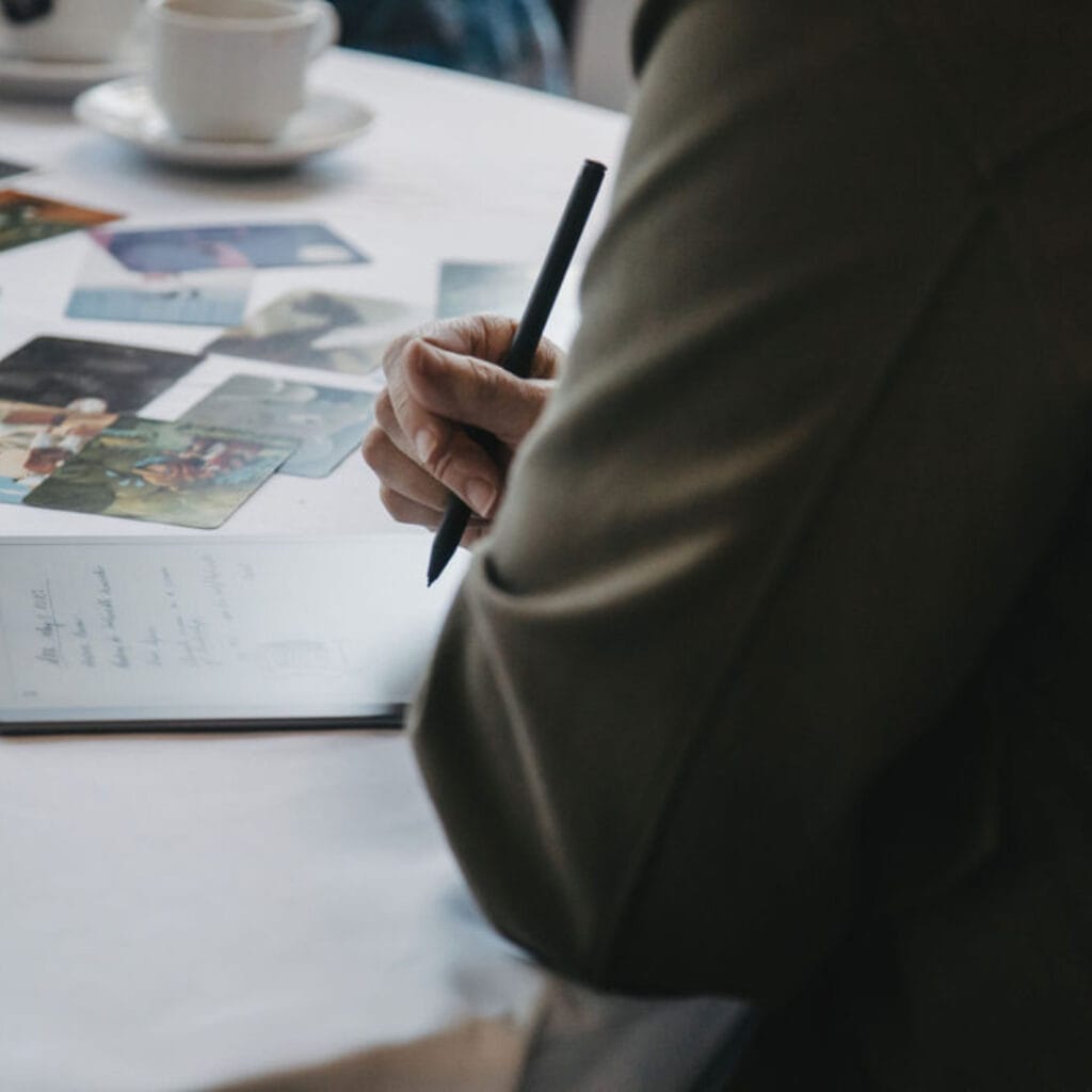 A person writes in a notebook with a pen at a table, surrounded by scattered photographs and a cup and saucer in the background.
