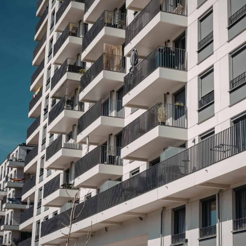 A modern apartment building with multiple floors, featuring white walls and black railings on each balcony, is thoughtfully maintained by housing providers. Several balconies have plants and other items, set against a clear, blue sky.
